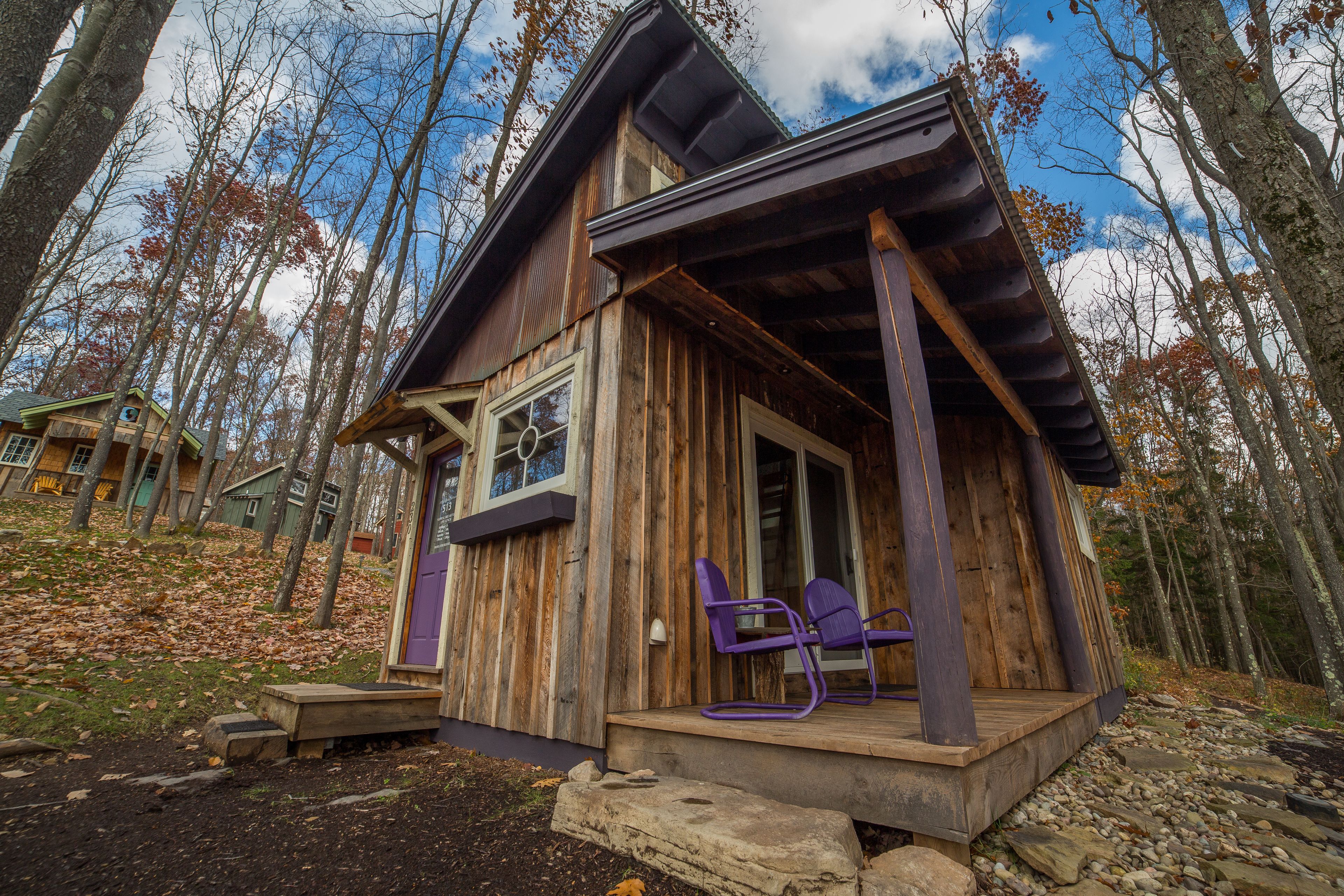 Una cabaña rústica con fachada de madera, un porche con sillas moradas y vistas al bosque circundante.