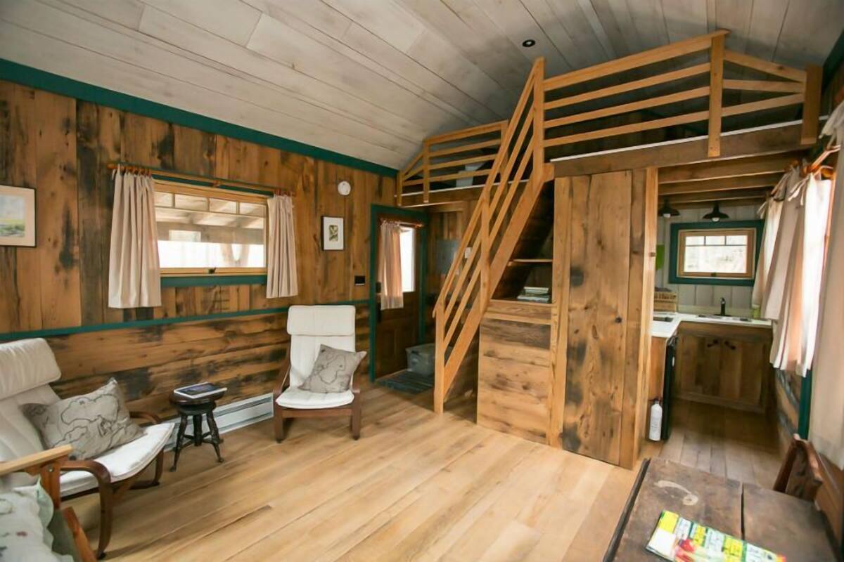 A cabin interior with a wooden floor, a staircase, a dining area, and a seating area with a sofa and chairs.