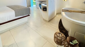 A modern bedroom with a large bed, a bathroom with a bathtub, and a view of a lush garden. - Pousada Ilha do Vento (São Miguel do Gostoso)