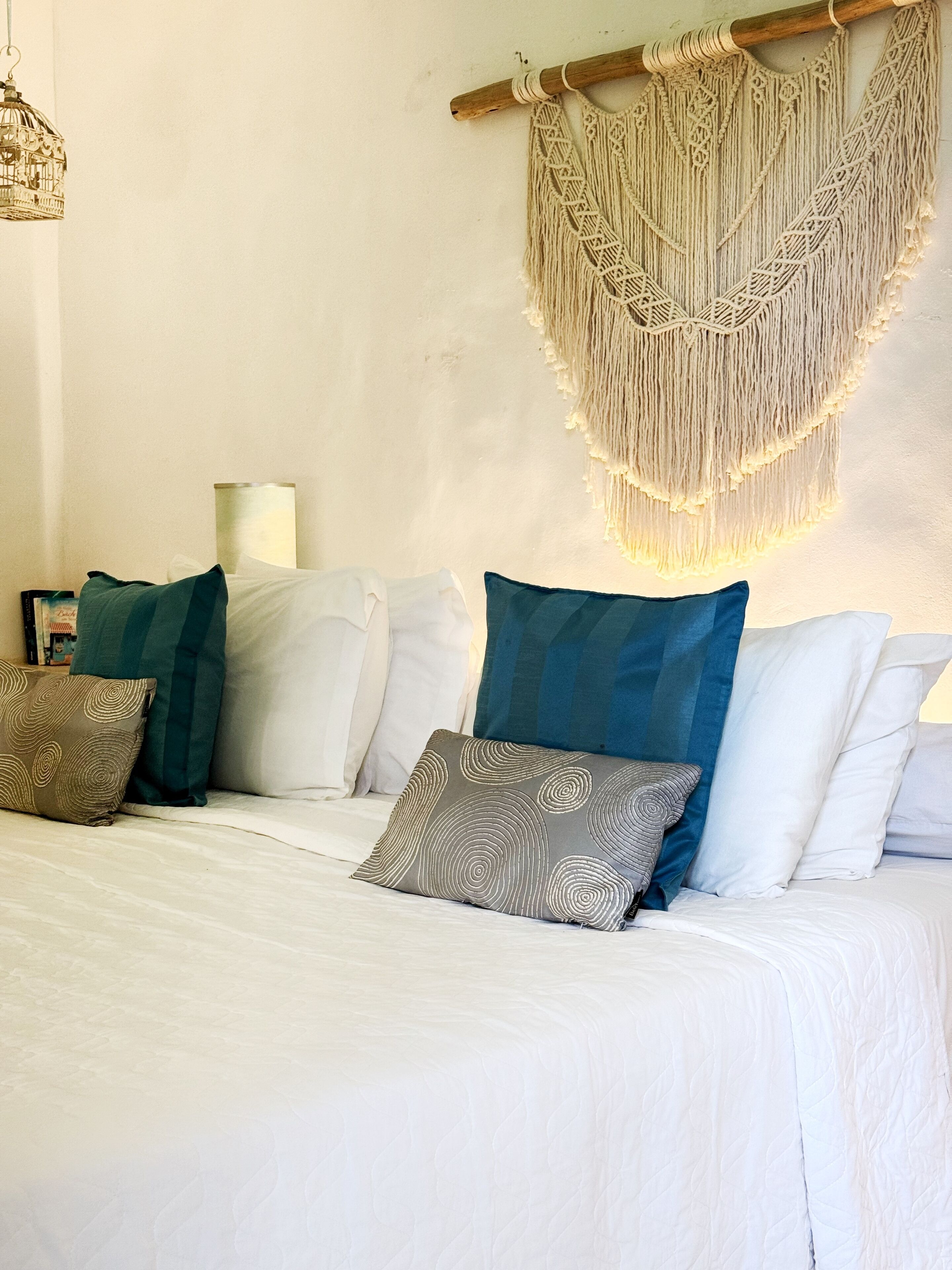 Ein Bett mit weißer Bettwäsche, mehreren Kissen in verschiedenen Farben und Mustern, sowie einer Wanddekoration aus Makramee.