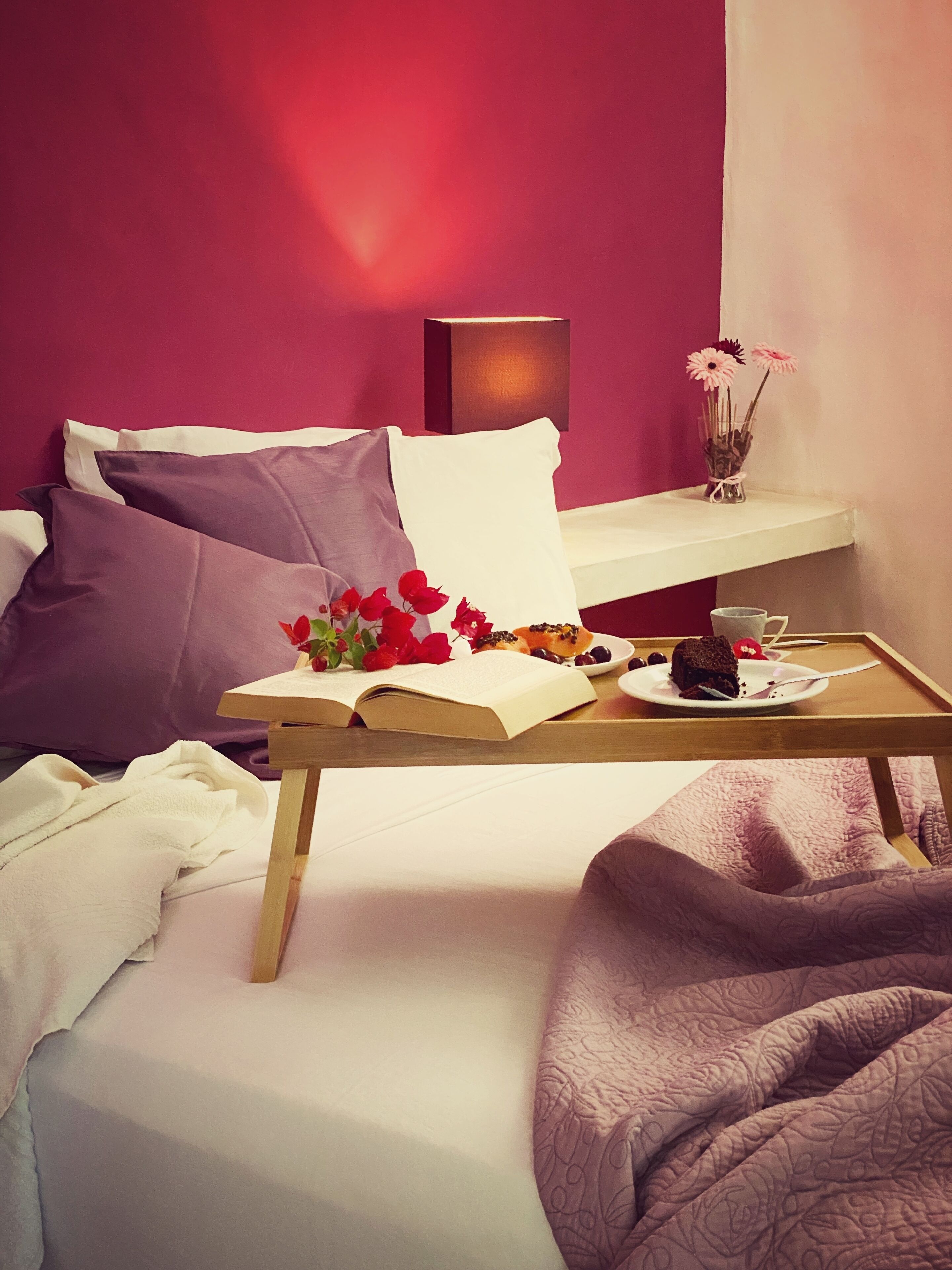 Un lit bien fait, agrémenté d’oreillers violets, une table de chevet avec un livre, des fleurs et une assiette contenant un dessert.