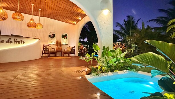 A modern outdoor lounge area with a wooden deck, a pool, and a seating area with a white wall and wooden ceiling. - Pousada Ilha do Vento (São Miguel do Gostoso)