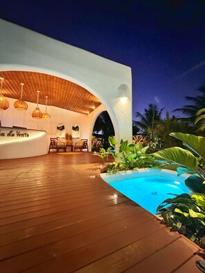 Un'area relax esterna moderna con un pontile in legno, una piscina e un angolo salotto con parete bianca e soffitto in legno.