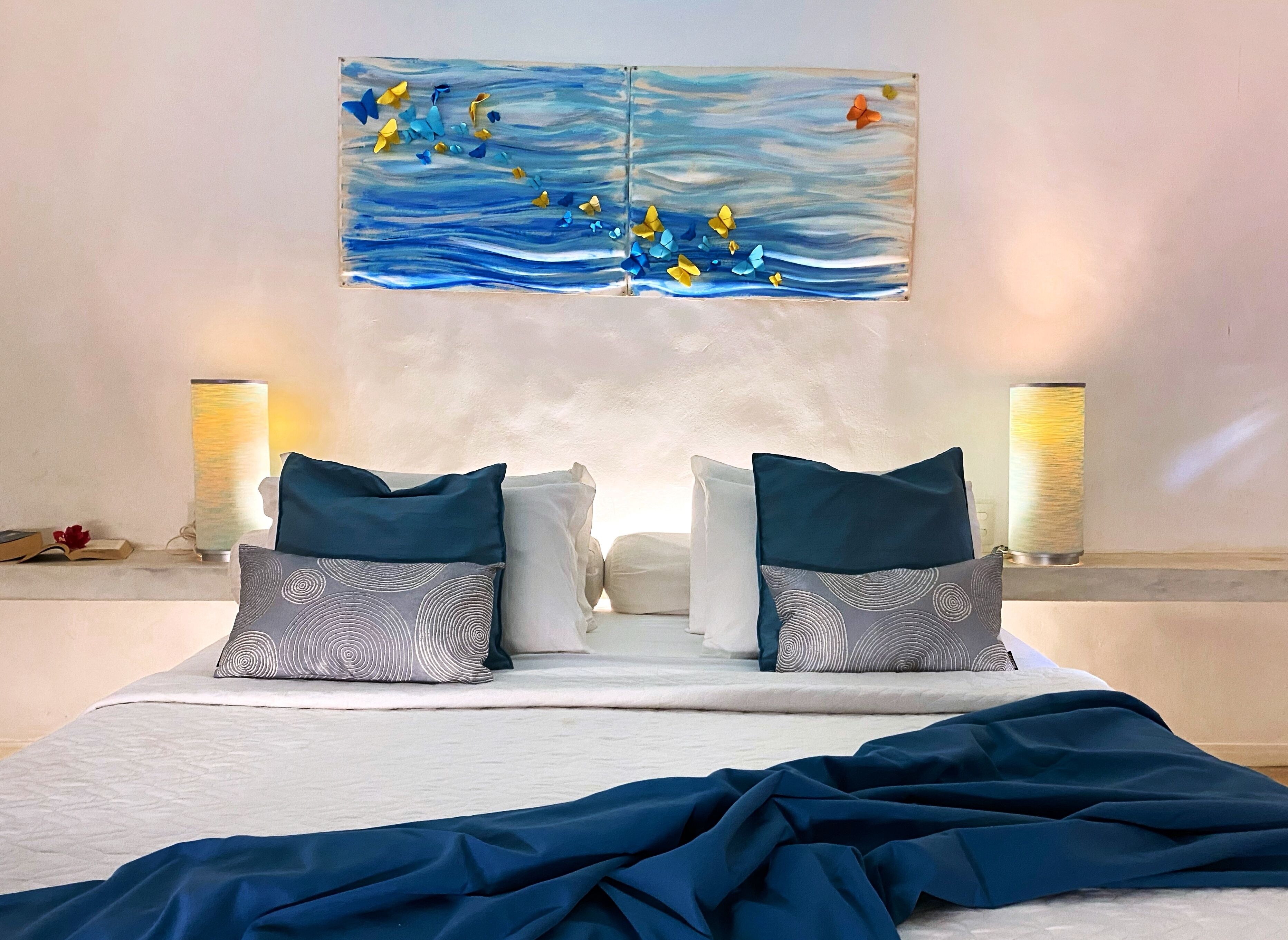 A bed with two pillows, a blue blanket, and a painting of butterflies on the wall.