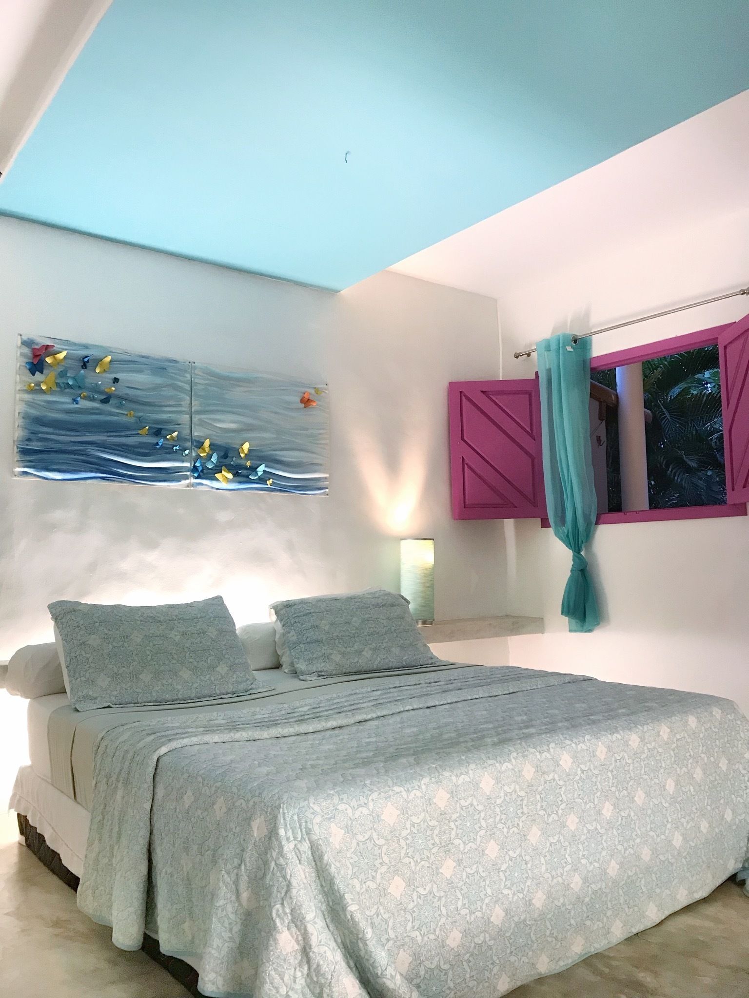 A bedroom with a bed, a painting, a window with curtains, and a wall-mounted light.