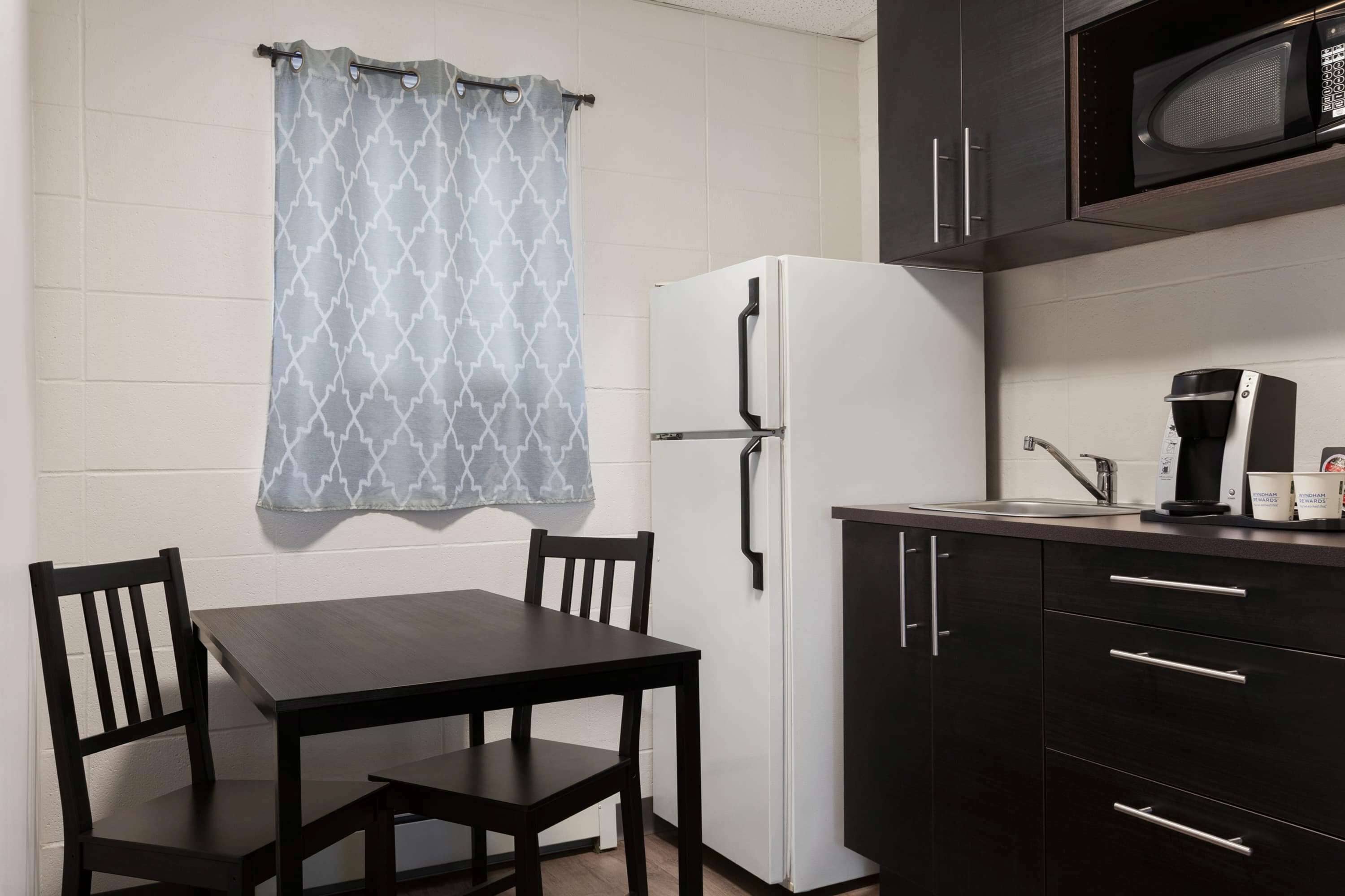 A compact kitchen with a white refrigerator, dark cabinets, a dining table with chairs, and a microwave above the counter.