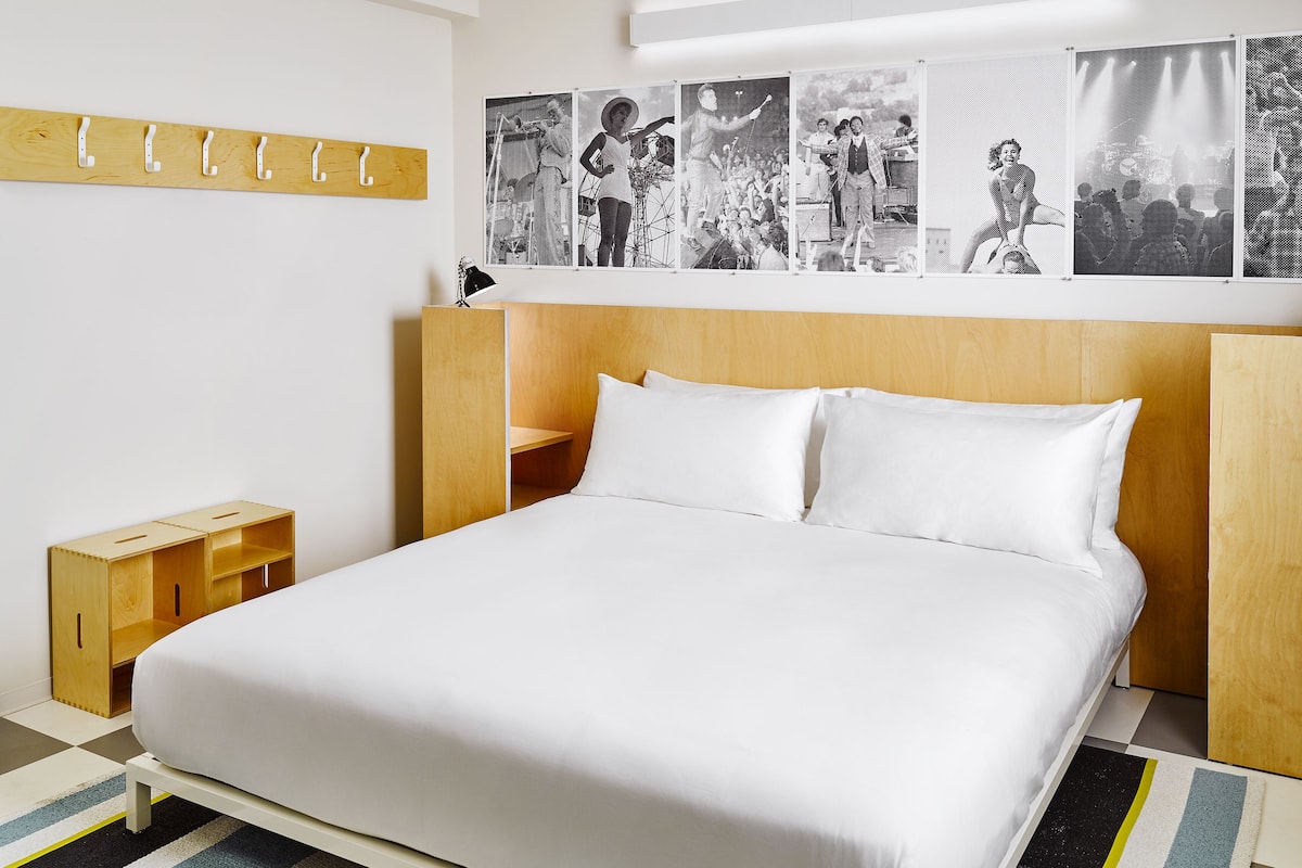 A modern bedroom with a wooden headboard, a bed with white linens, a small wooden nightstand, and a wall-mounted coat rack.