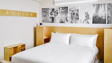 A modern bedroom with a wooden headboard, a bed with white linens, a small wooden nightstand, and a wall-mounted coat rack.