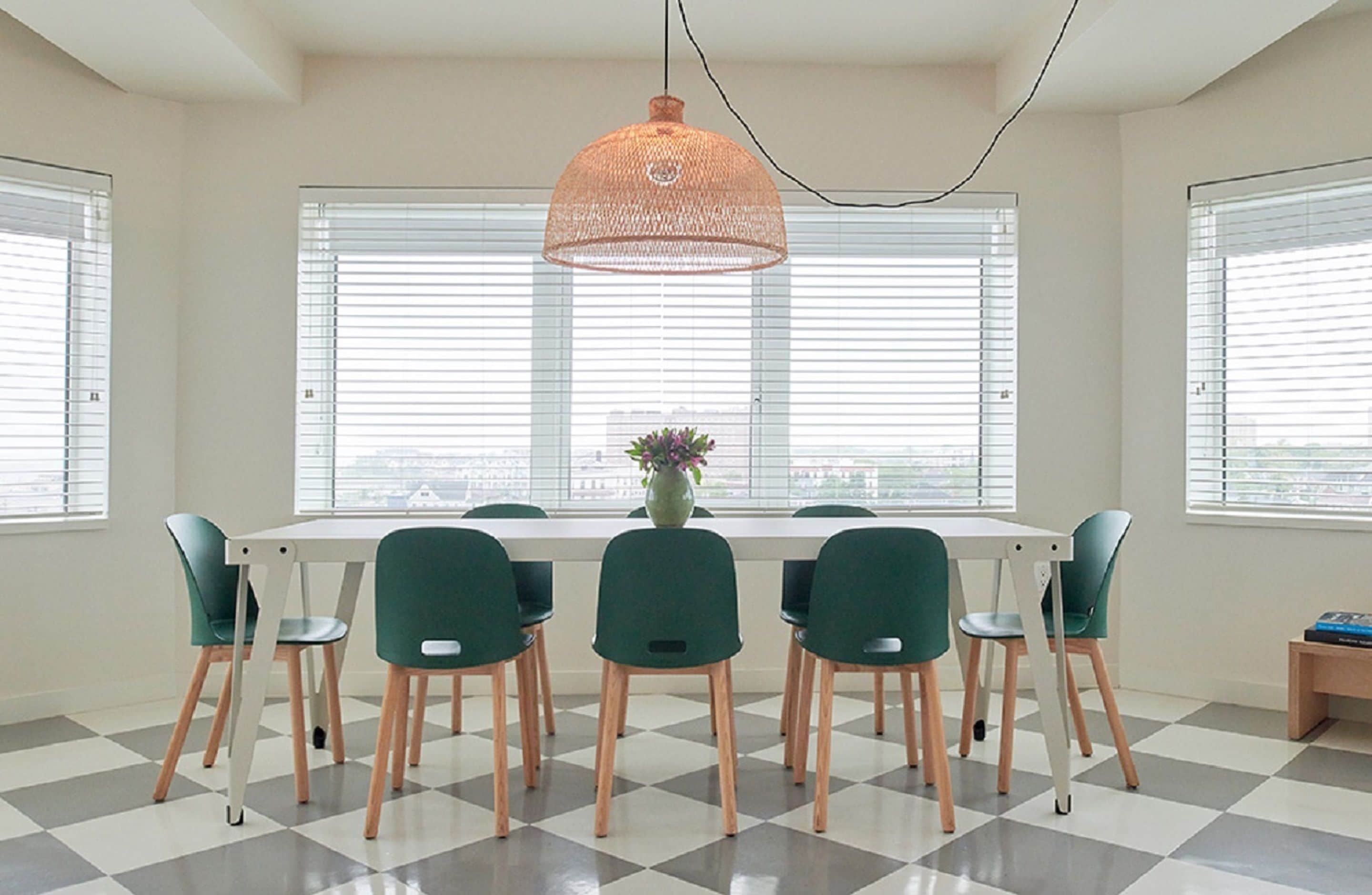 A dining area with a white table, green chairs, a woven pendant light, and a vase with flowers.