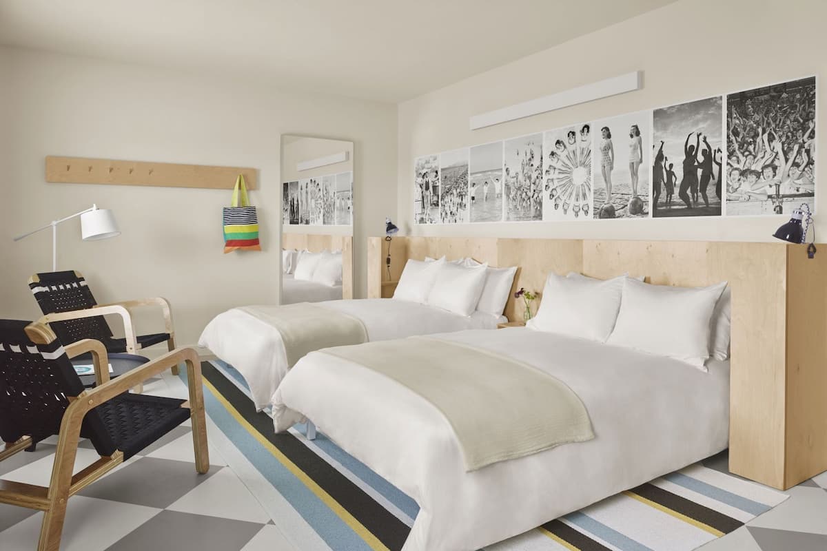 A hotel room with two beds, a desk, a chair, a lamp, and a wall with black and white photographs.