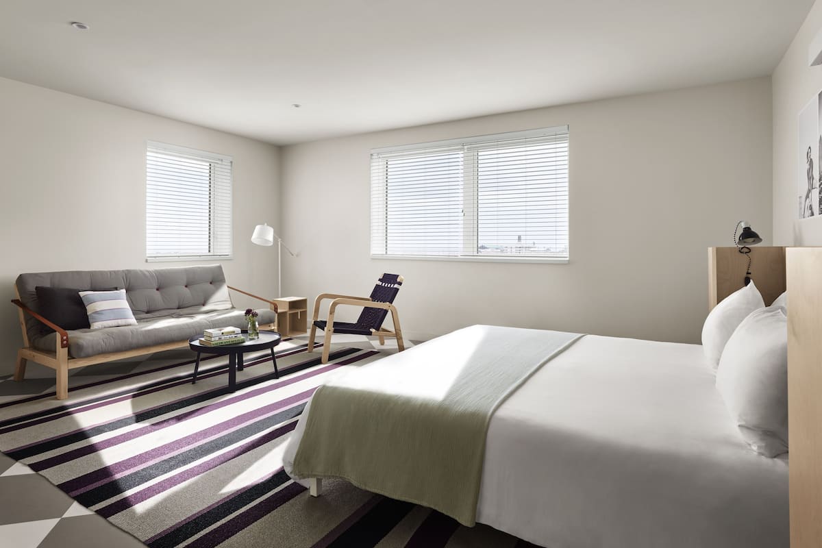 A modern hotel room with a bed, sofa, armchair, and a small table.