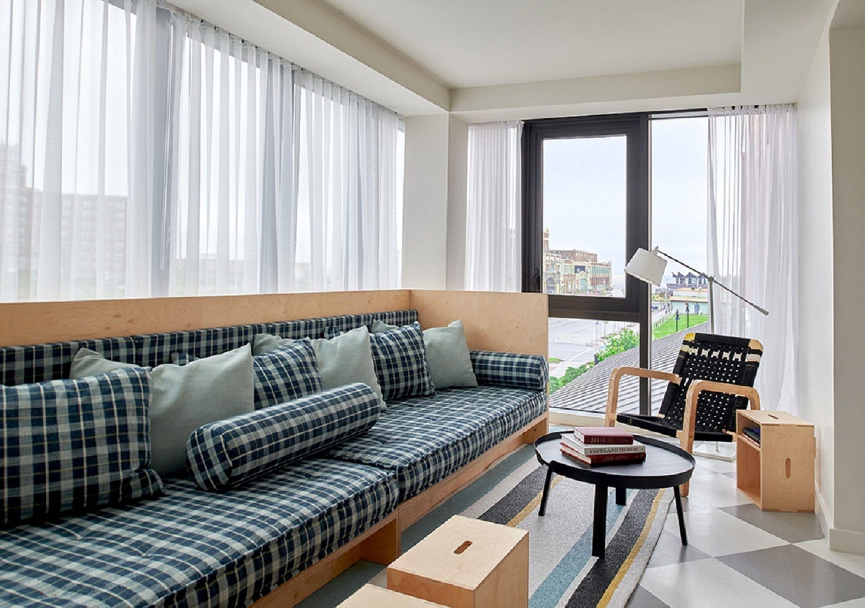 A modern living room with a sofa, a small table, and a chair. There are large windows with curtains, a view of buildings outside, and a lamp on the table.