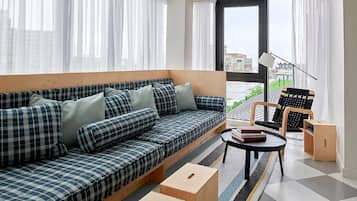 A modern living room with a sofa, a small table, and a chair. There are large windows with curtains, a view of buildings outside, and a lamp on the table.