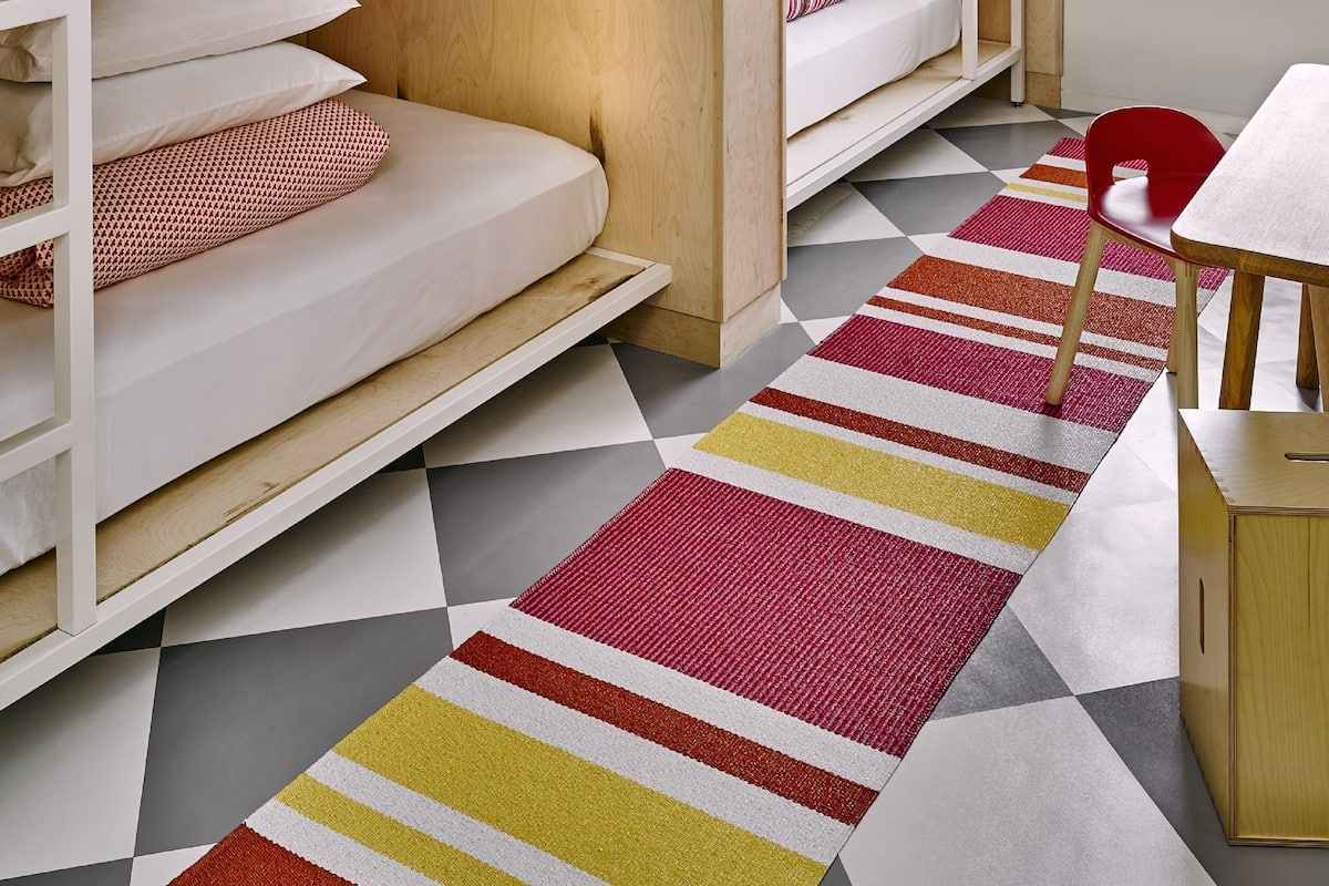 A dormitory room with bunk beds, a small table, and a colorful striped rug.
