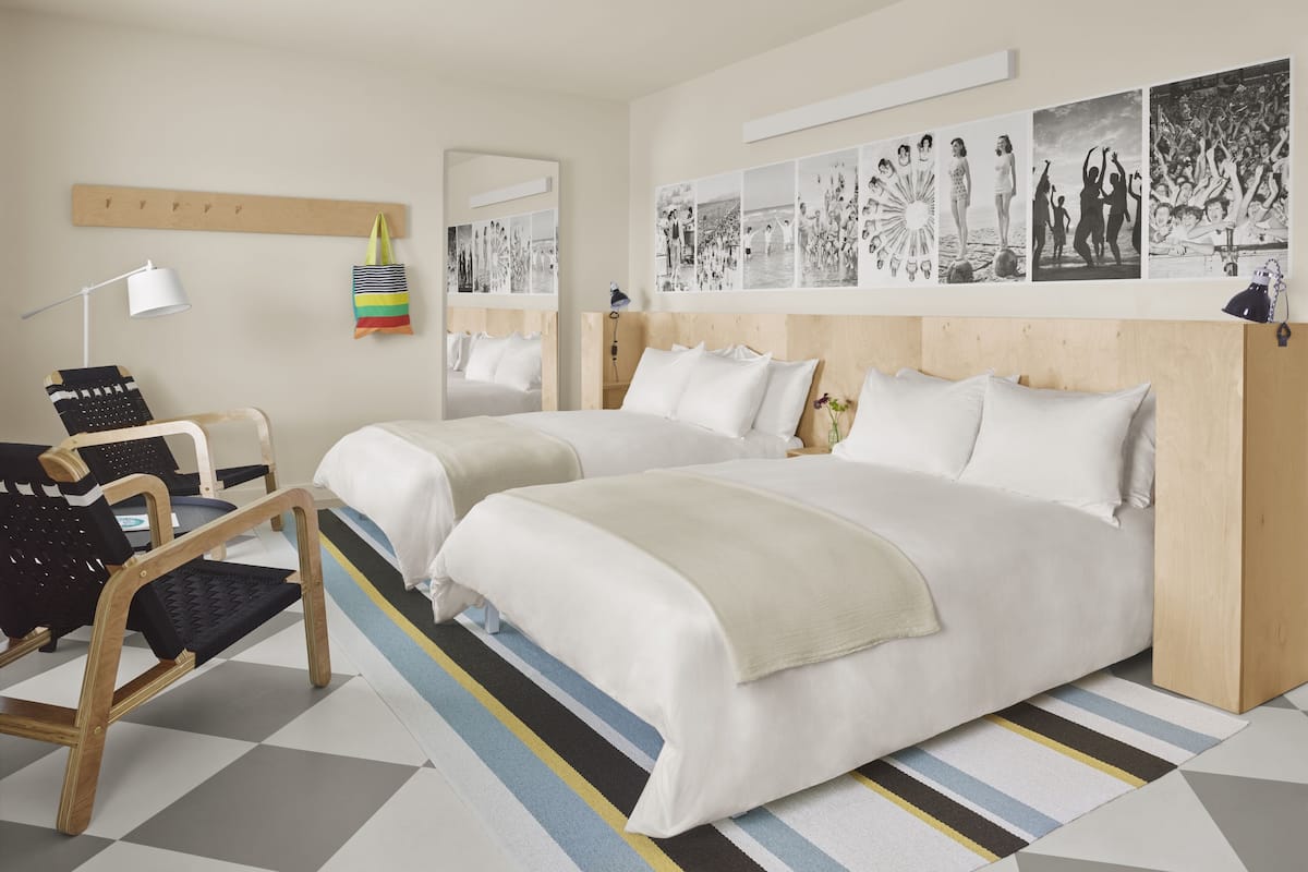A hotel room with two beds, a desk, and a chair. There are wall-mounted pictures and a mirror.