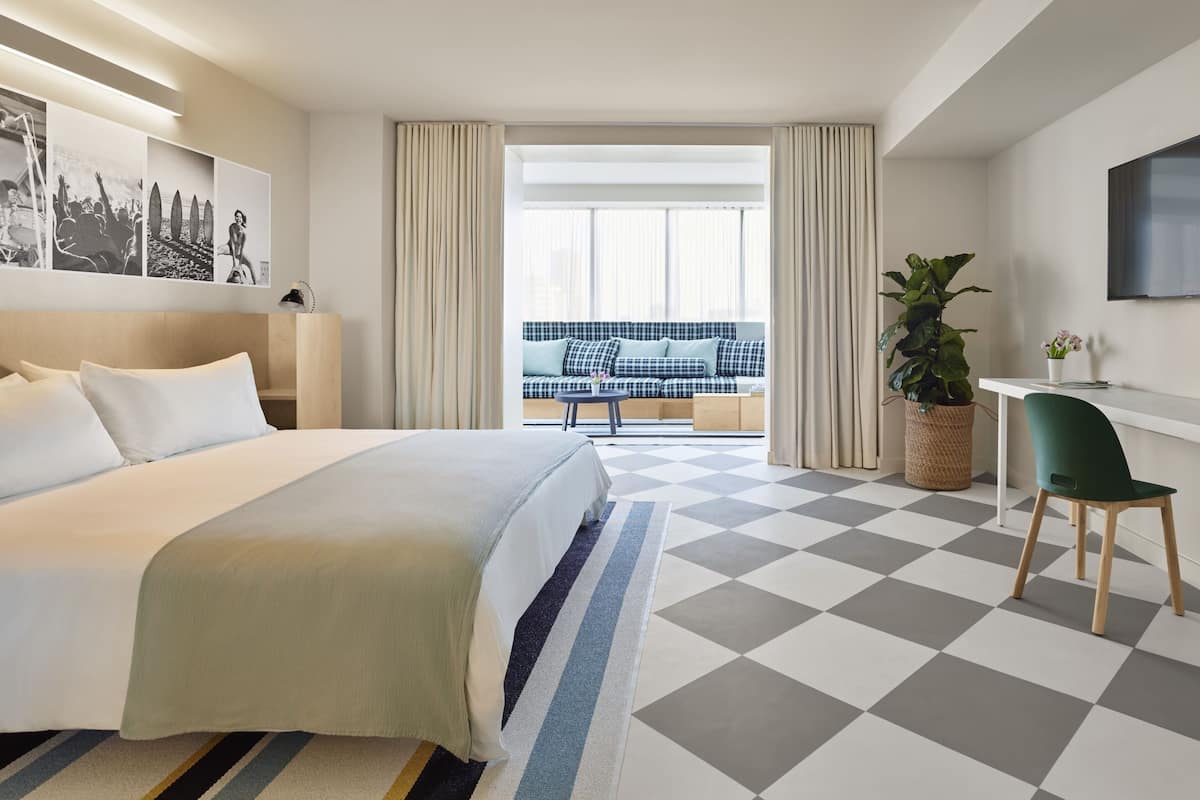 A hotel room with a large bed, a desk, a chair, a potted plant, and a balcony with a sofa.