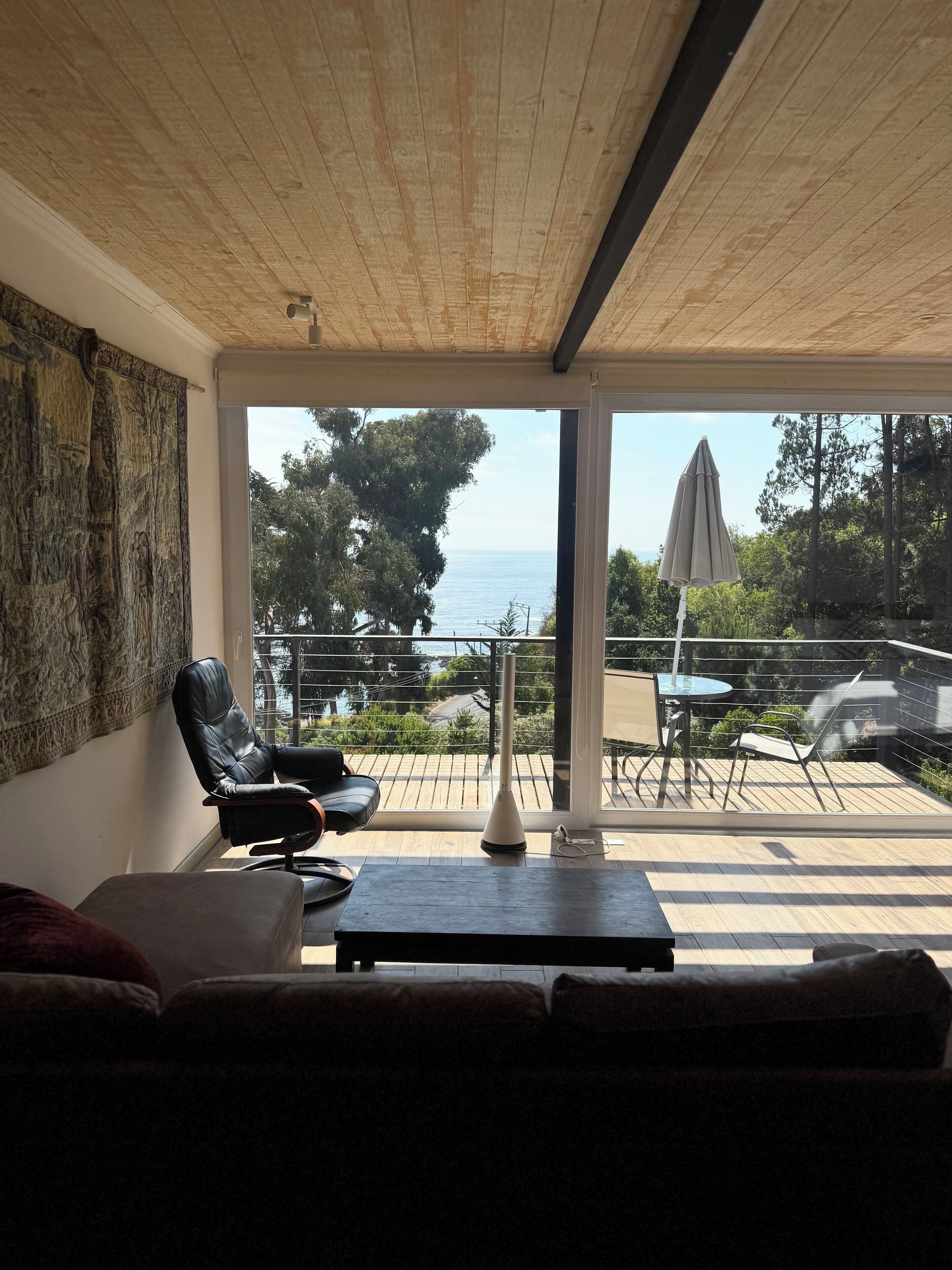 A modern living room with a wooden ceiling, a large window, and a view of the sea. There is a black leather chair, a white sofa, and a small table with a white vase.