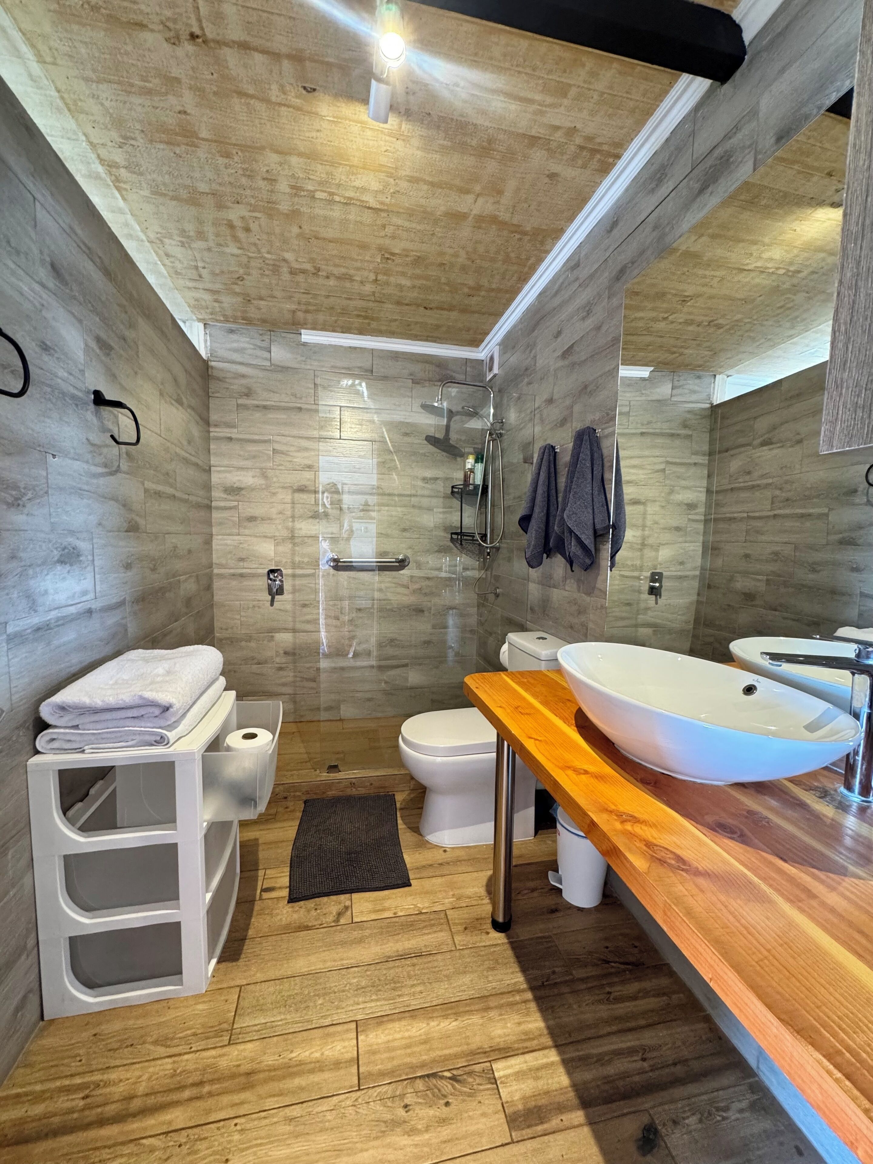 A modern bathroom with a wooden floor, a large white sink, a toilet, and a shower area.