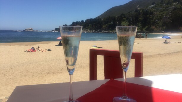 Zwei Sektgläser auf einem Tisch mit roter Servette, mit Blick auf einen Strand mit Menschen und Sonnenschirmen.