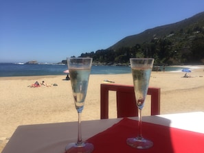 Dos copas de champán sobre una mesa con un mantel rojo, con vista a una playa con gente y sombrillas.