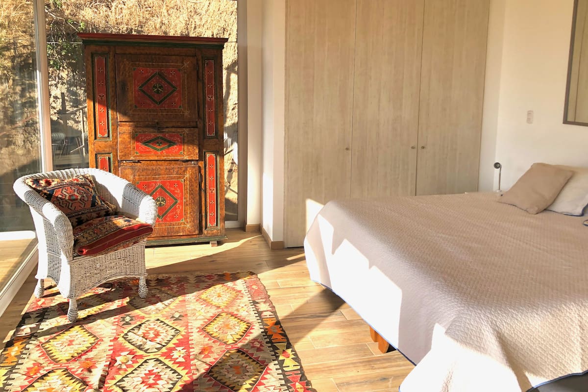 Une chambre avec un grand lit, une chaise en osier avec un coussin à motifs, une armoire en bois ornée de panneaux décoratifs et un tapis à motifs sur le sol.