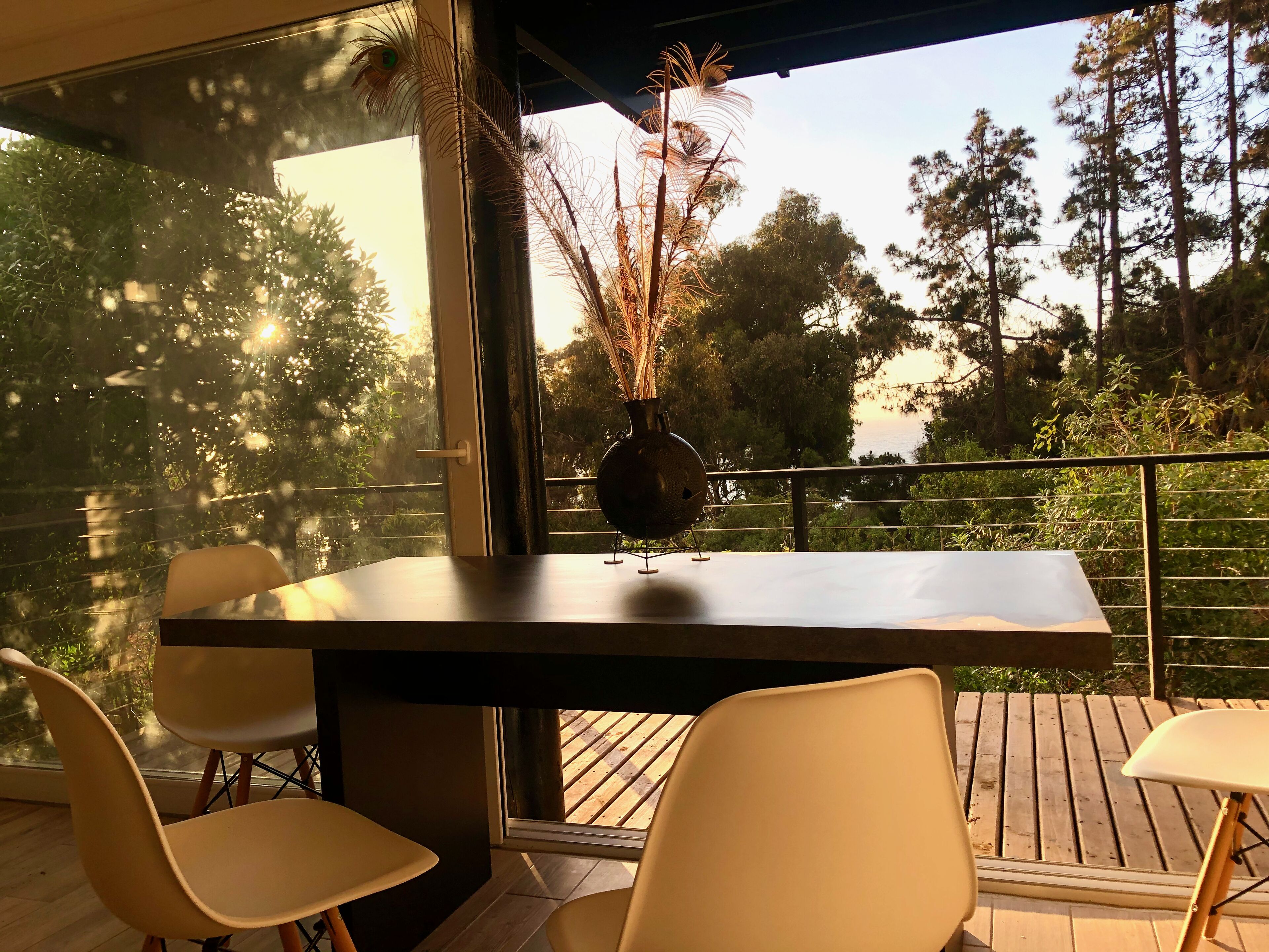 Un comedor moderno con una mesa y sillas, un jarrón con plantas secas, y vistas a árboles y un cuerpo de agua a través de grandes ventanales.
