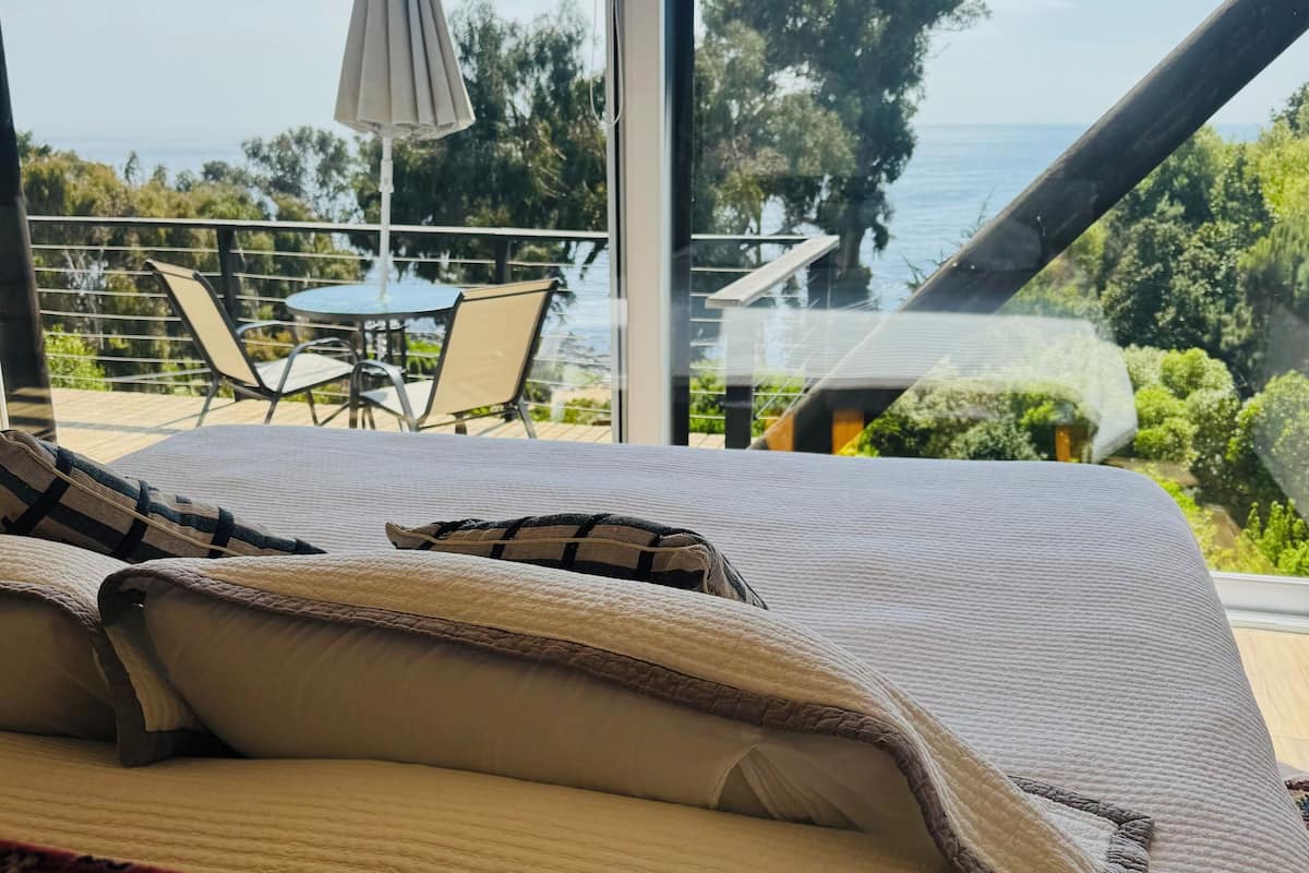 Une chambre dotée d’une grande fenêtre donnant sur un balcon aménagé avec du mobilier d’extérieur et offrant une vue sur la mer.