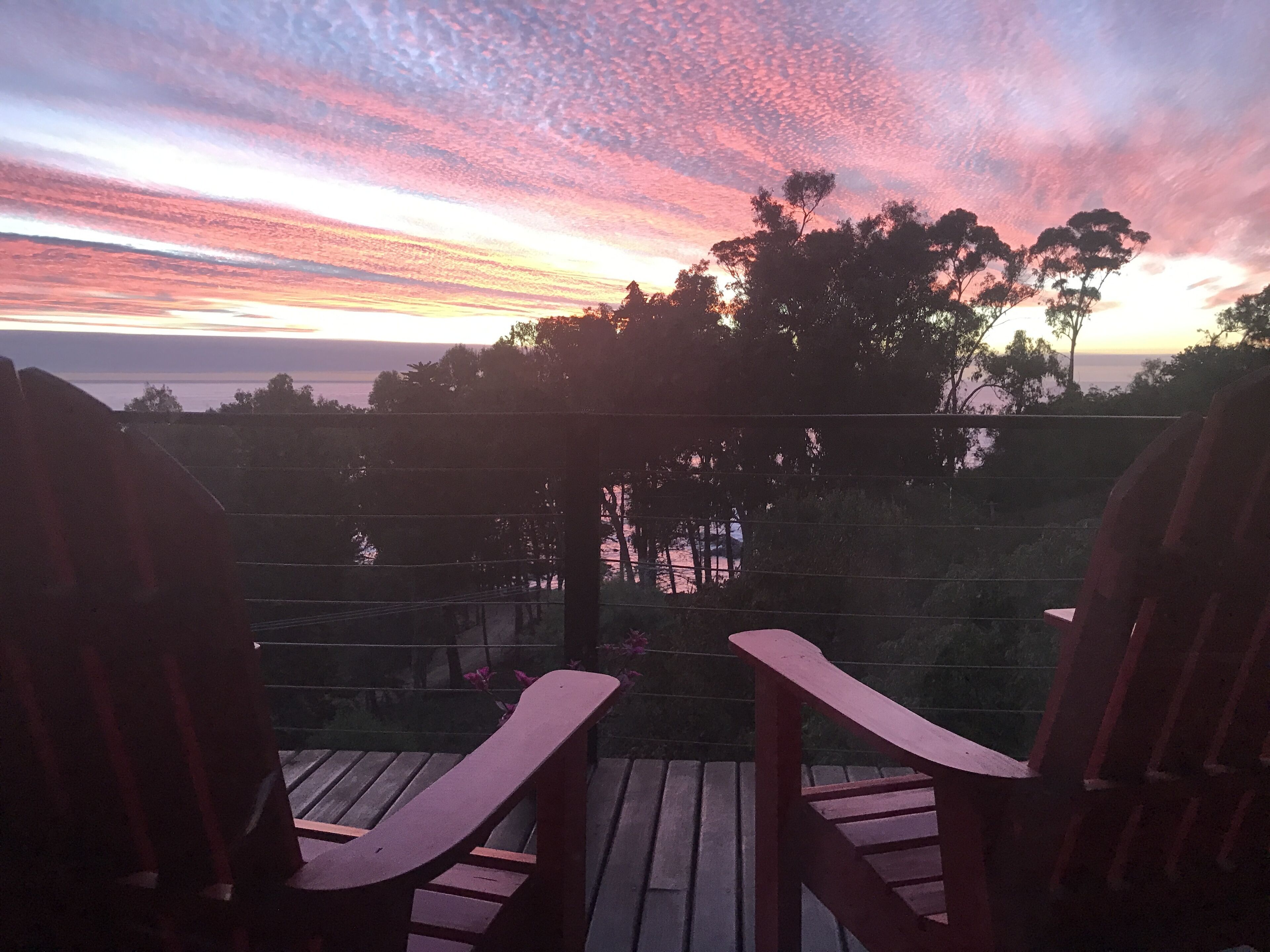 Une terrasse en bois avec deux fauteuils Adirondack donnant sur un coucher de soleil au-dessus d’une étendue d’eau.