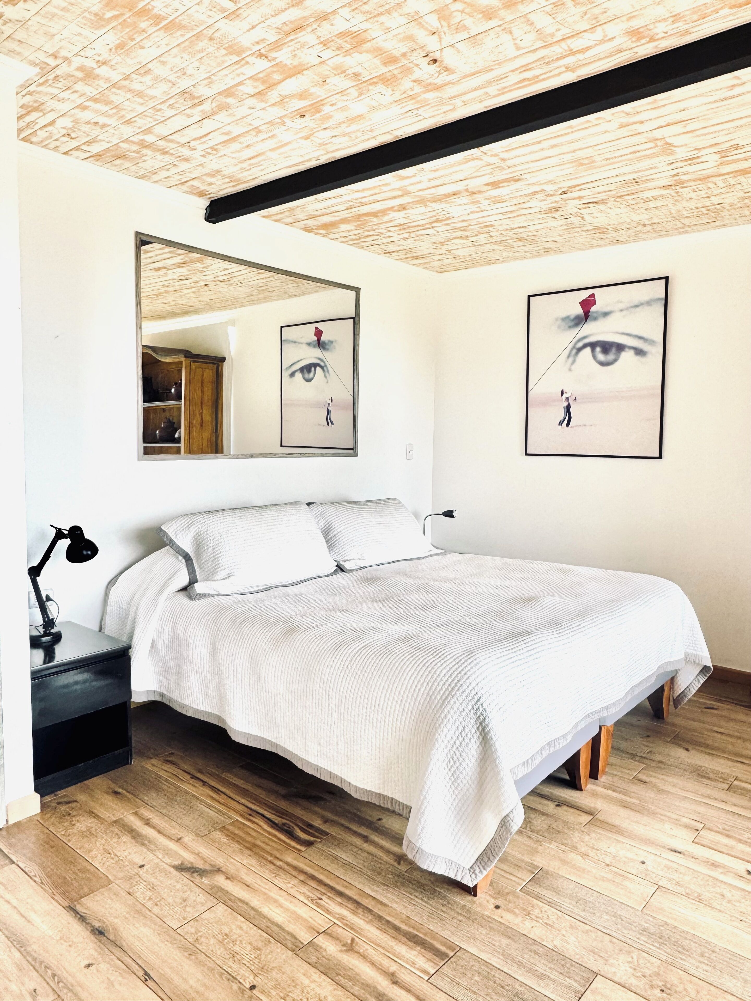 A bedroom with a wooden ceiling, a bed with white bedding, a nightstand with a lamp, and two framed artworks on the wall.