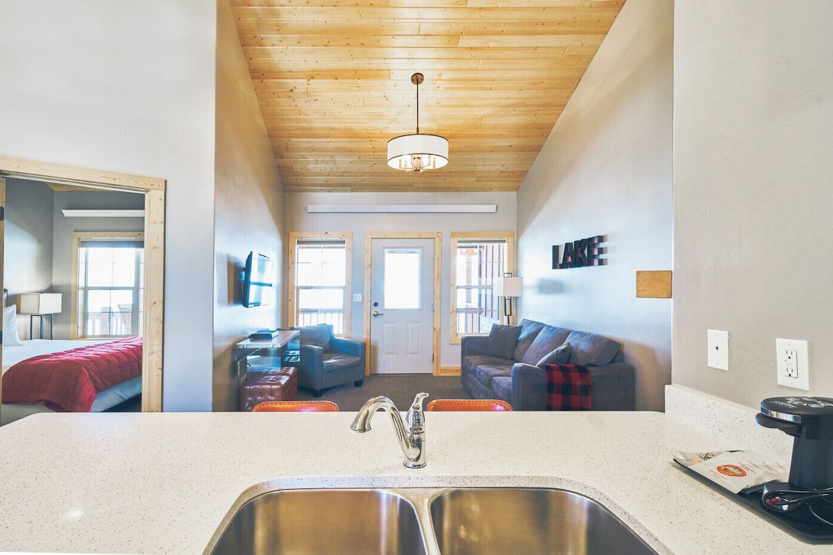 A compact kitchen with a double sink, a dining area with a table and chairs, and a living room with a couch and a TV.