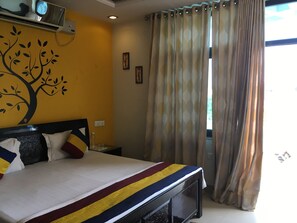 A hotel room with a bed, a wall-mounted air conditioner, a window with curtains, and a wall decoration.