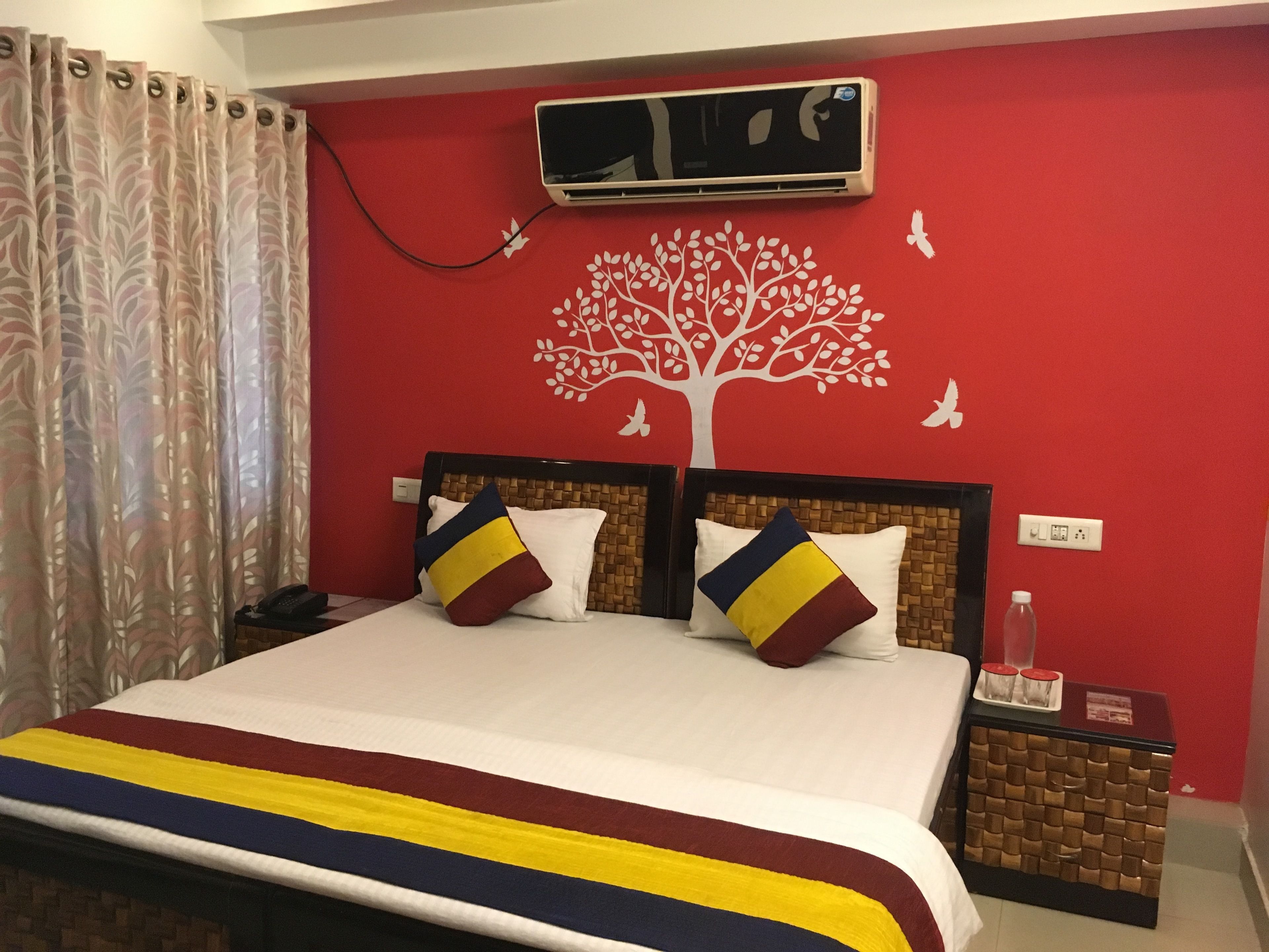 A hotel room with a large bed, two pillows, a nightstand, an air conditioning unit, and a wall with a tree decal.