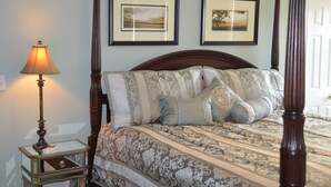 A four-poster bed with a patterned bedspread and multiple pillows, a bedside table with a lamp, and two framed pictures on the wall.