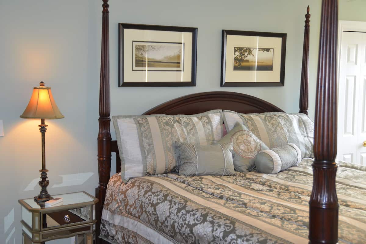 A four-poster bed with a patterned bedspread and multiple pillows, a bedside table with a lamp, and two framed pictures on the wall.