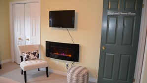 A room with a fireplace, a television mounted on the wall, a door labeled "Harp and Crown Room," a chair with a floral cushion, and a small ottoman.