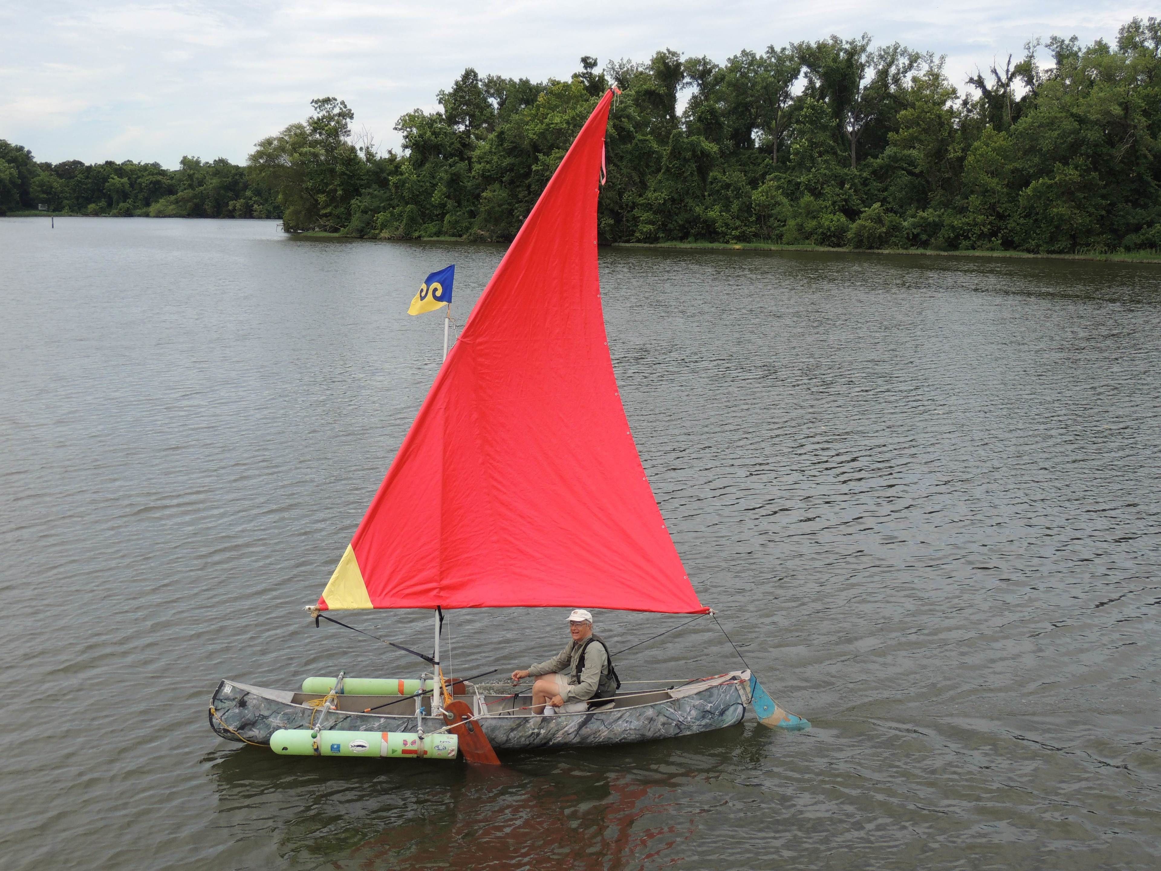 Un petit bateau à voile rouge sur une étendue d’eau calme, entouré d’arbres.