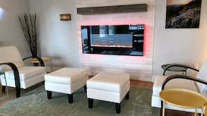 Ein Wohnzimmer mit Flachbildfernseher an einer Ziegelwand, zwei weißen Ledersofas, einem kleinen runden Tisch und einem grünen Teppich.