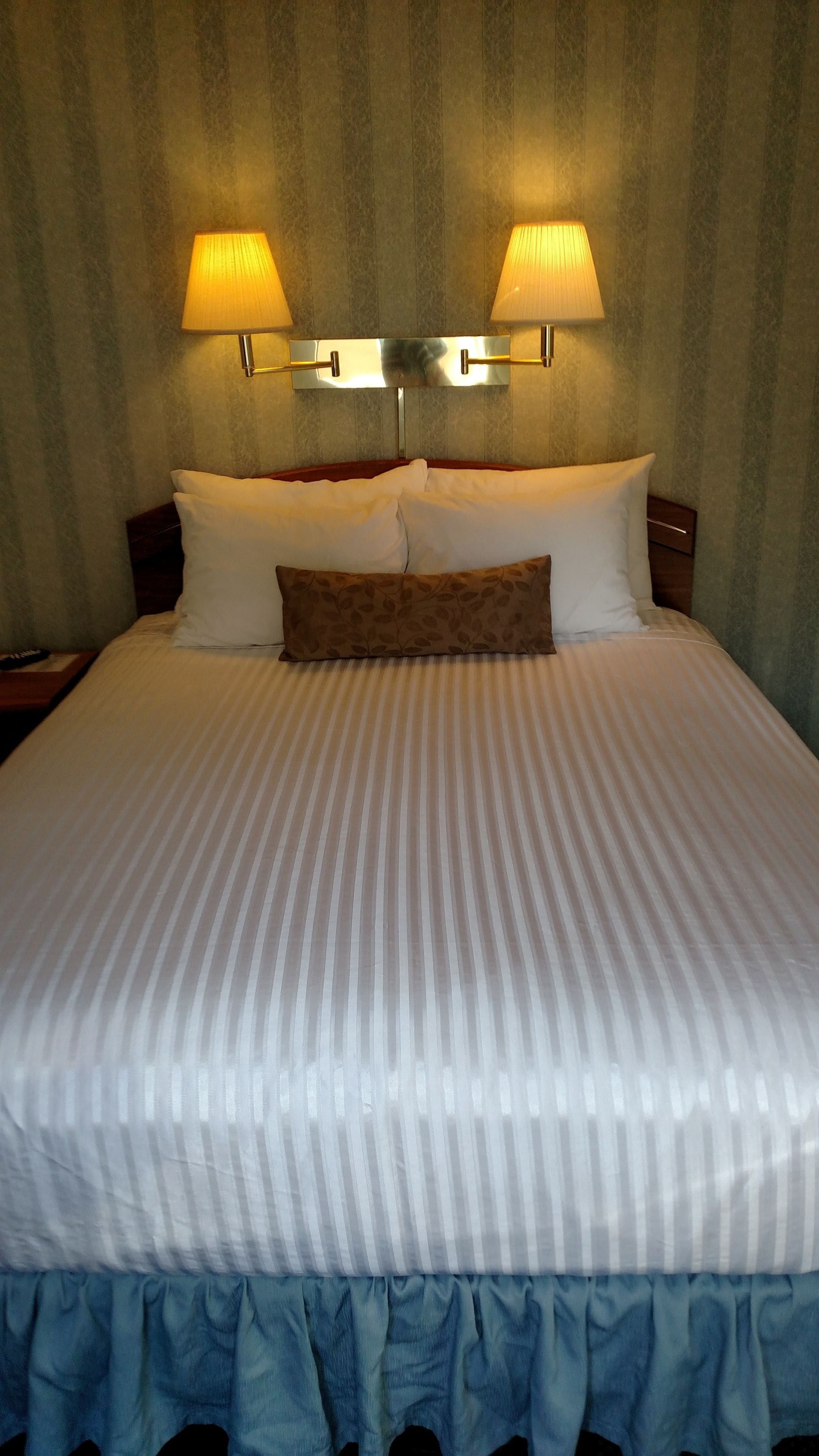 A neatly made bed with white and striped bedding, two wall-mounted lamps, and a patterned pillow.