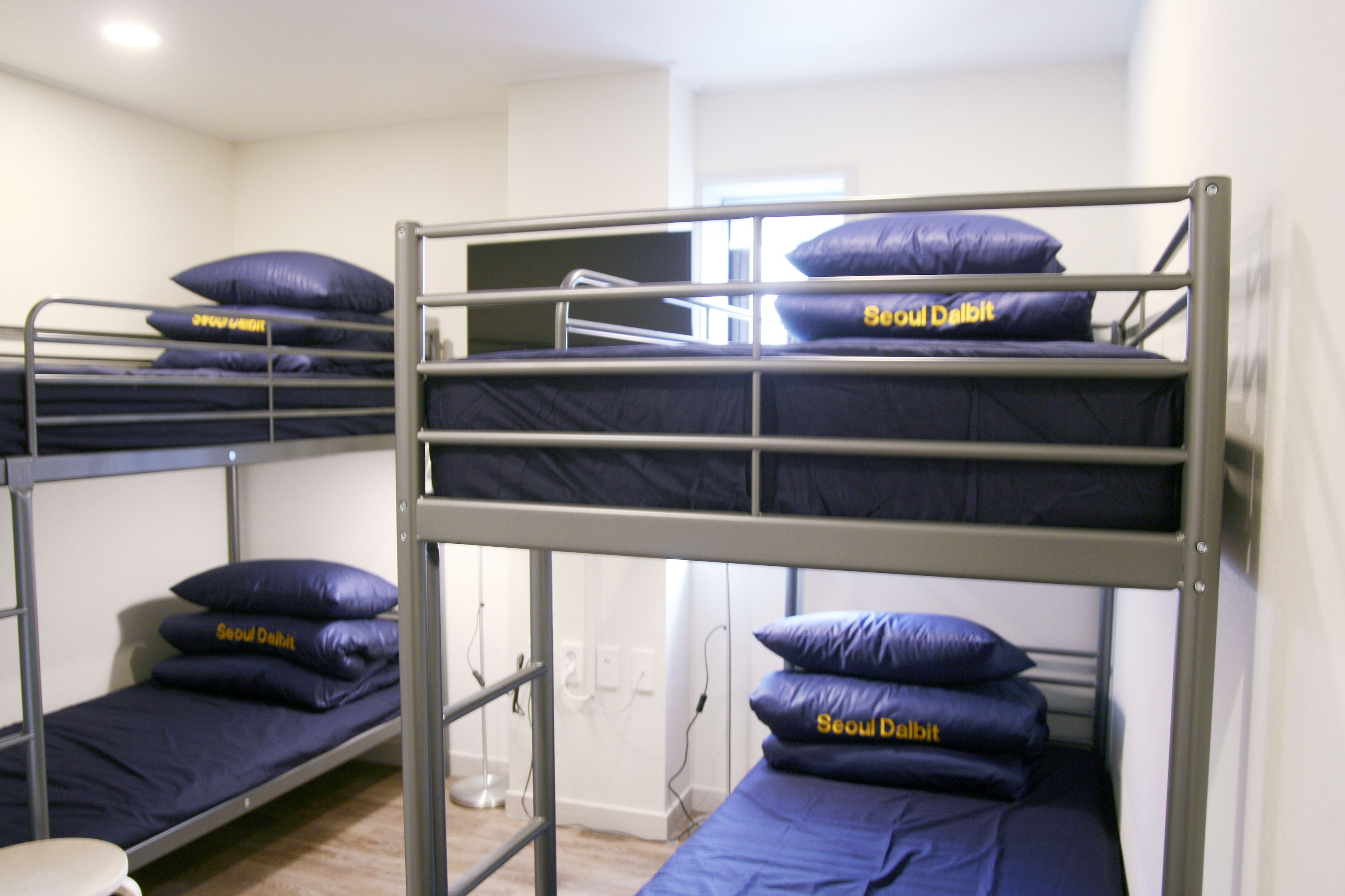 A dormitory room with bunk beds, each with a blue mattress and pillows labeled "Seoul Dalbit".
