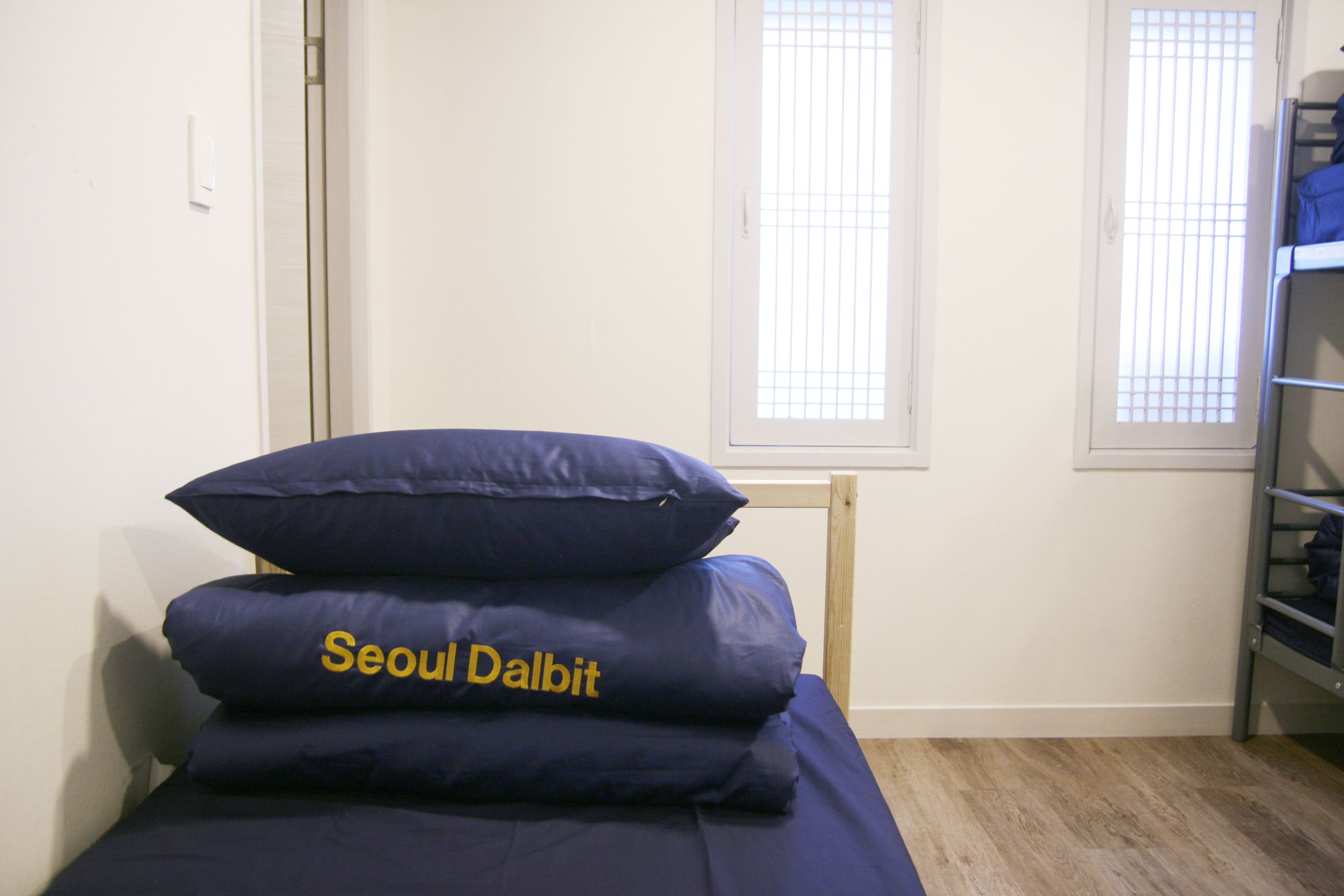 A single bed with blue bedding and a pillow labeled "Seoul Dalbit" in a room with white walls and a window.