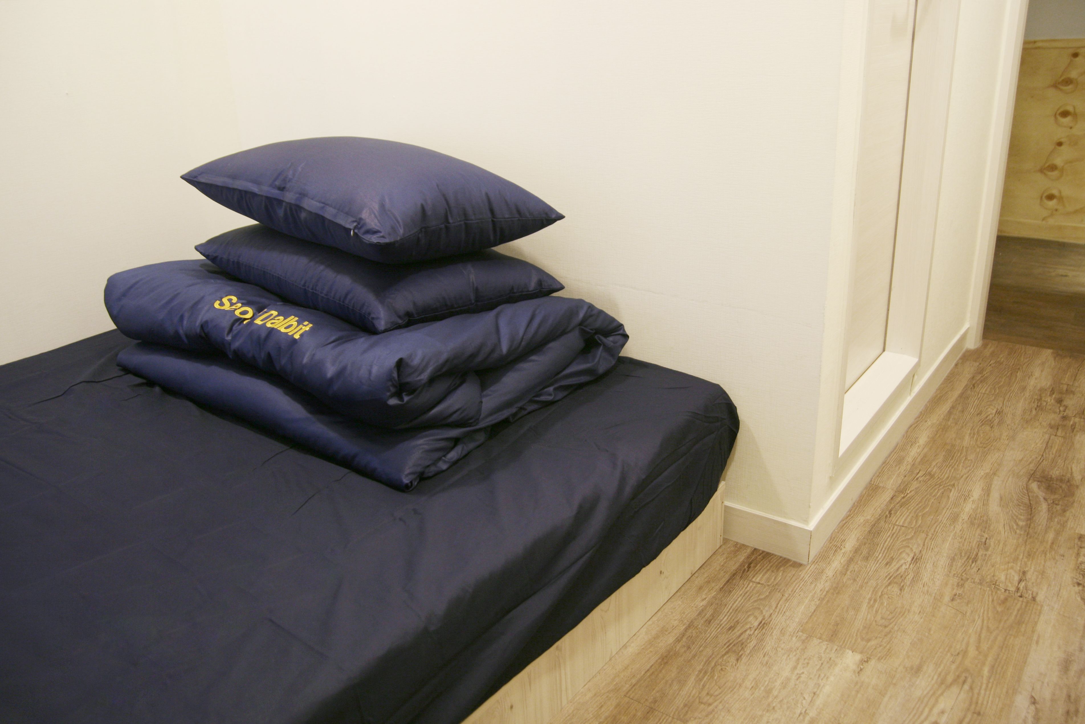 A bed with dark blue bedding and pillows, a wooden floor, and a white wall.