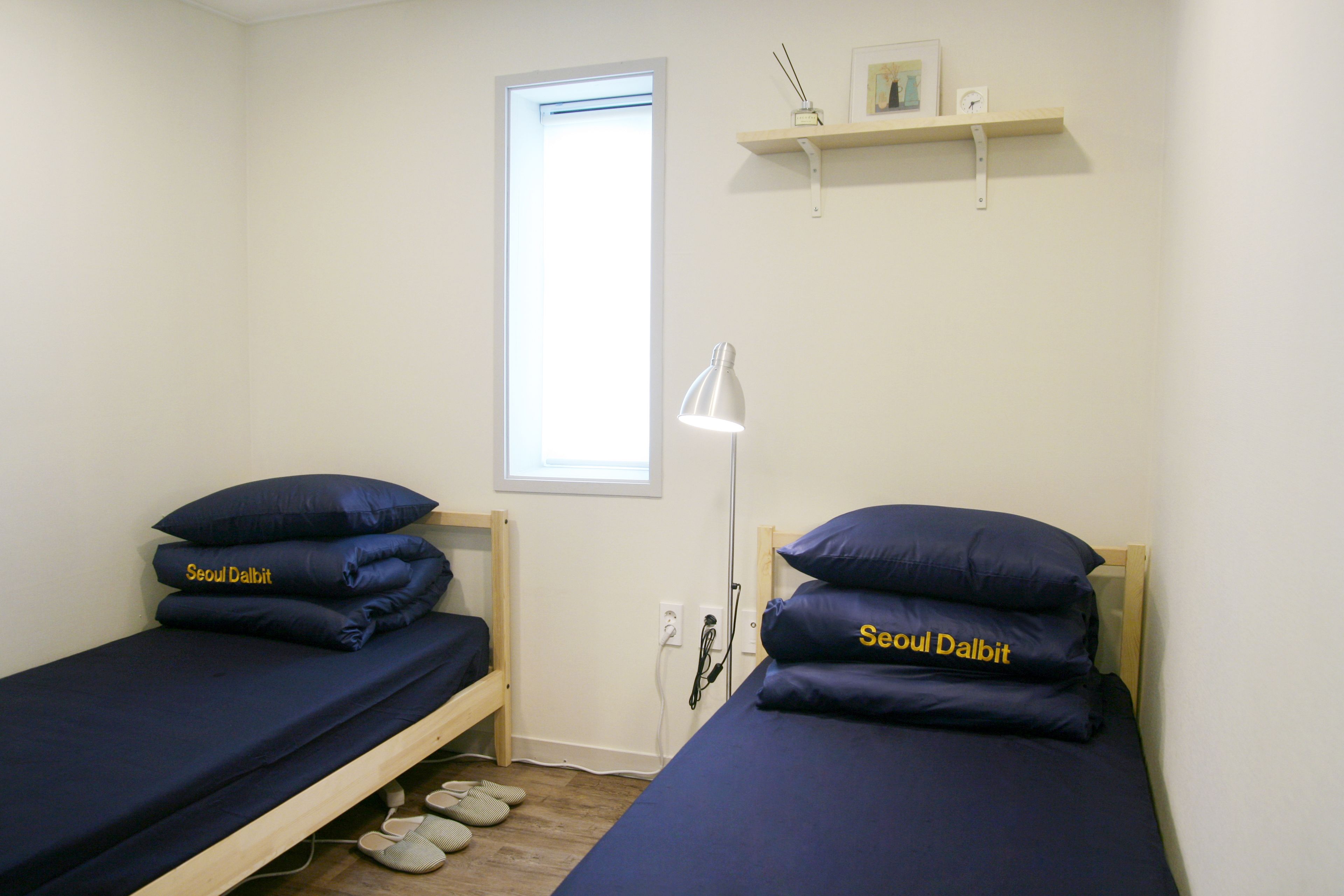 A small, clean room with two single beds, each with dark blue bedding and pillows labeled "Seoul Dalbit." There's a window providing natural light, a white desk lamp, and a small shelf above the beds.