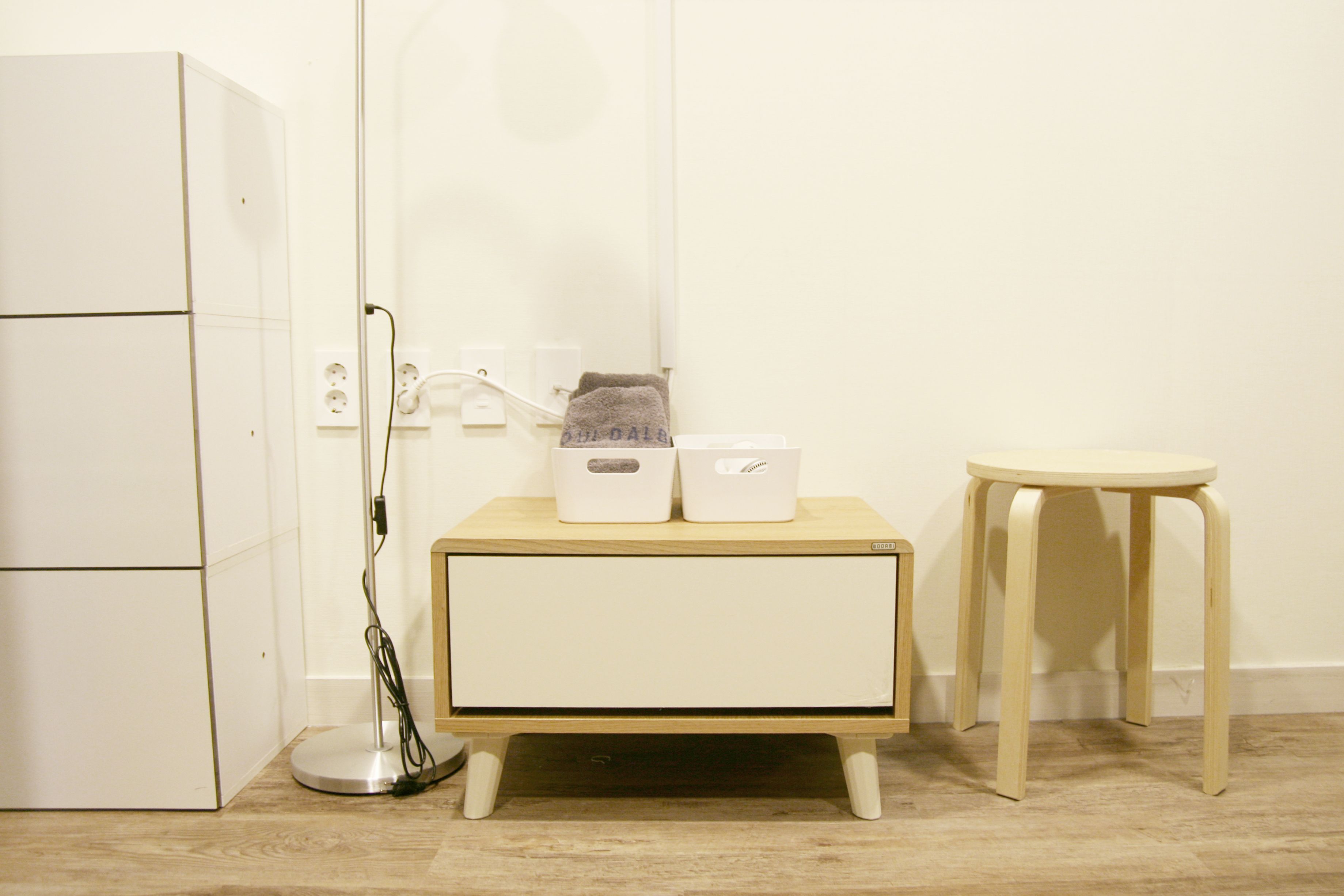 A small, clean room with a white wardrobe, a bedside table with two boxes, a lamp, and a stool.