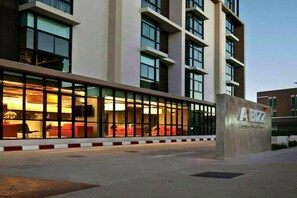 A modern hotel with a glass facade, a visible lobby area with seating, and a signboard with the hotel's name.