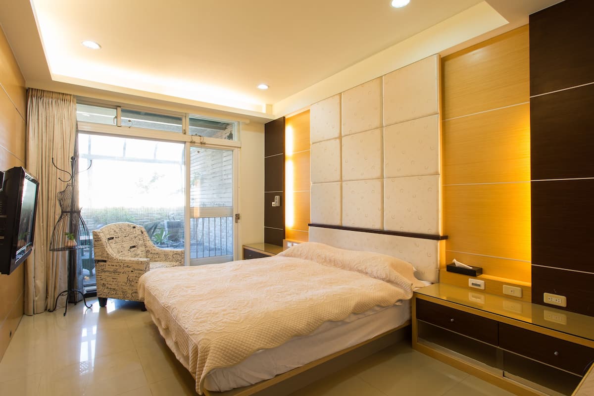 Quarto moderno com uma cama grande, porta de vidro que dá para uma varanda e área de estar com uma cadeira.