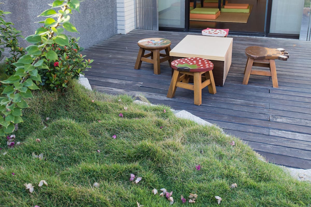 Um pátio com mesa e bancos de madeira, um pequeno jardim com grama e flores, e uma porta de vidro deslizante que dá para um espaço interior com almofadas.