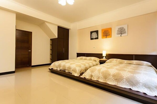 A bedroom with two beds, a nightstand, and a wall-mounted lamp. There are three framed pictures on the wall.