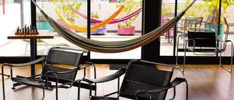 A room with a hammock, two black chairs, a chessboard, and a view of buildings through large windows.