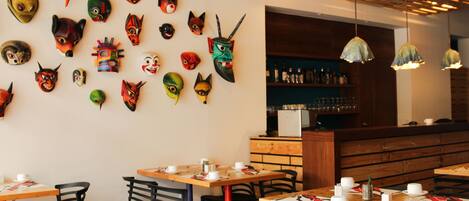 A restaurant with wooden tables and chairs, a wall of colorful masks, and a bar area with bottles.