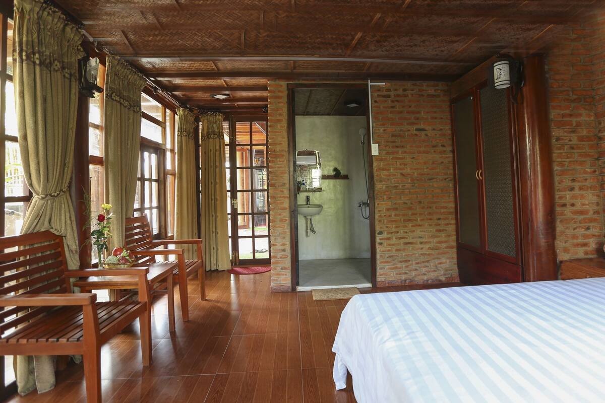A room with a wooden floor, a bed, a wooden bench, a table with a vase of flowers, and a bathroom with a shower and sink.